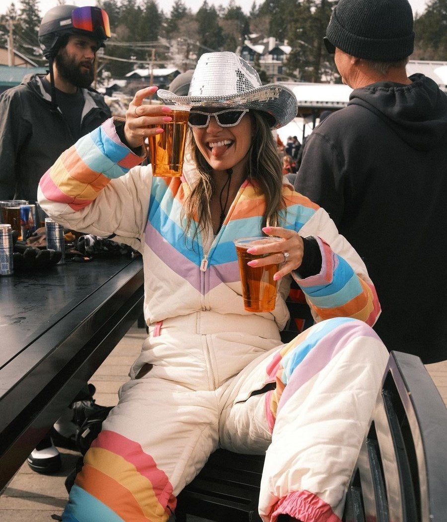 Woman in a rainbow retro ski suit and sequin cowboy hat toasting with beers at a ski lodge