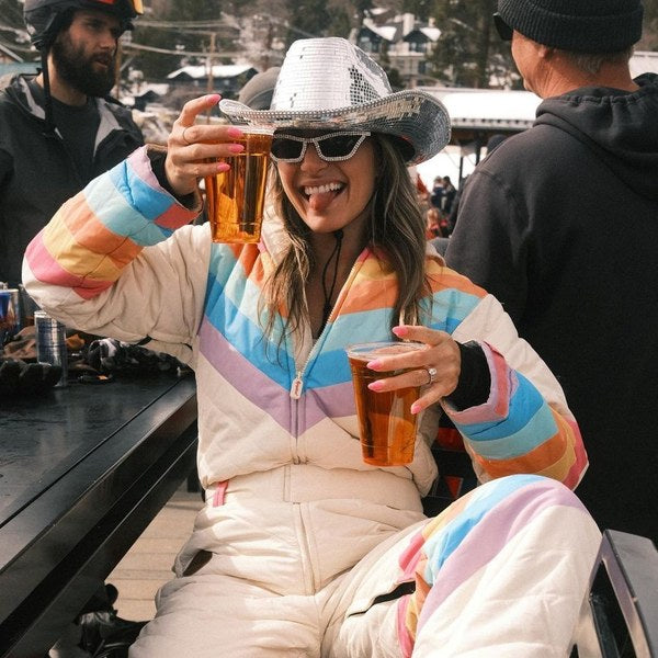 Woman in a rainbow retro ski suit and sequin cowboy hat toasting with beers at a ski lodge