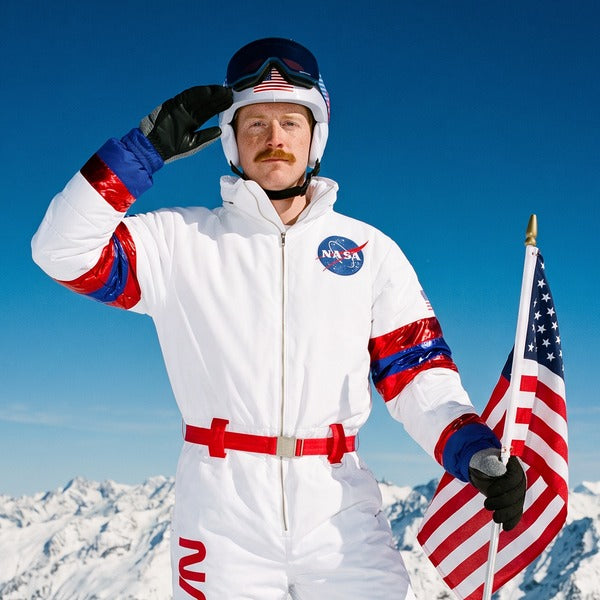 Man in a white NASA men's snow suit saluting with an American flag on a snowy mountain