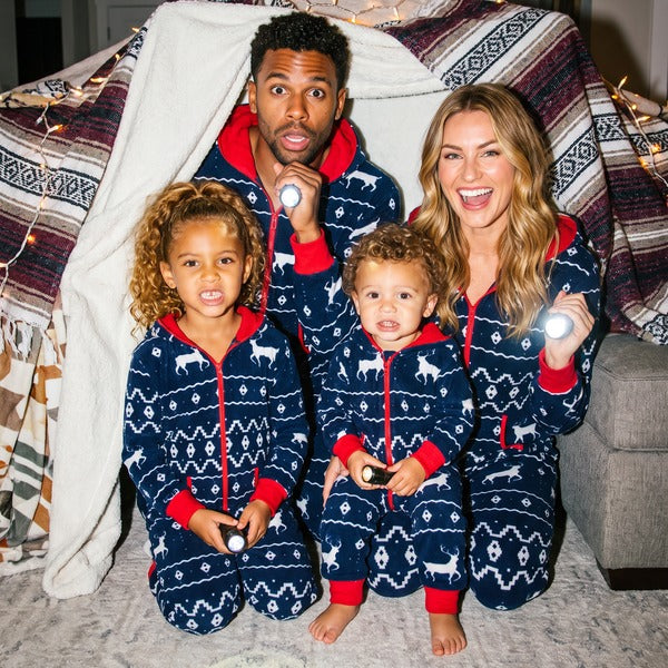 Family in matching navy Fair Isle reindeer Christmas jumpsuits with flashlights in a blanket fort